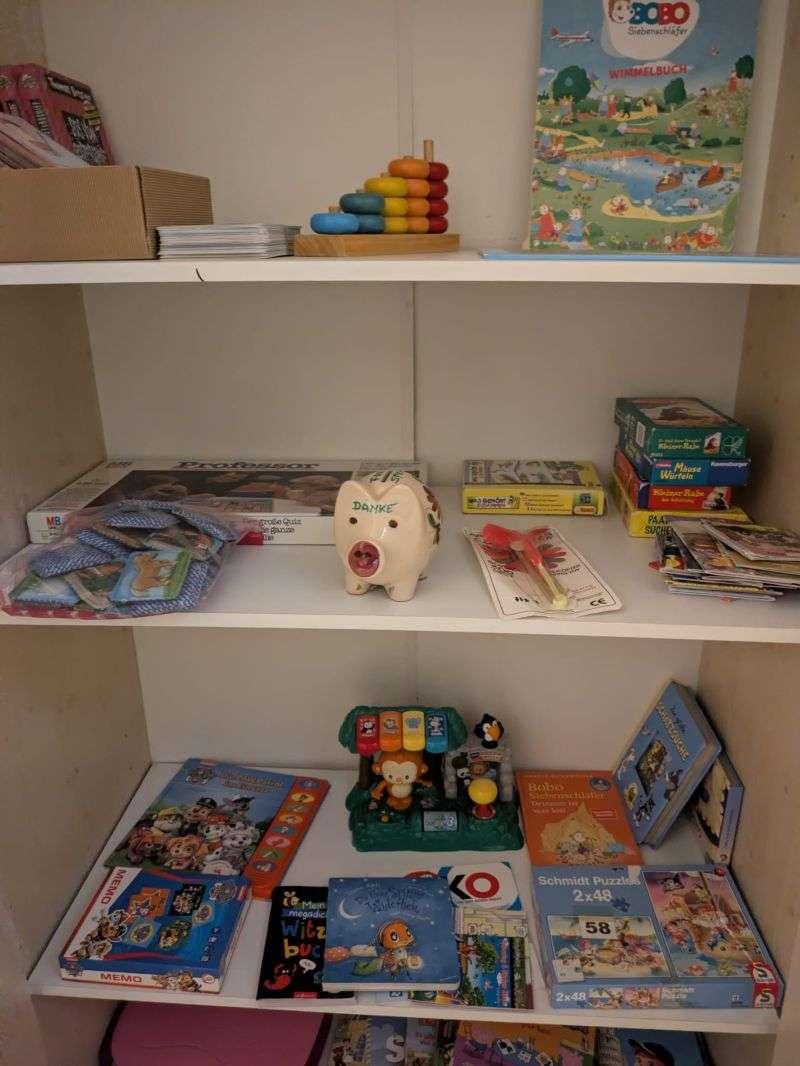 Ein Regal mit verschiedenen Kinderbüchern, Spielen und Spielsachen – Inhalt des Spielzeug-Tauschkastens.