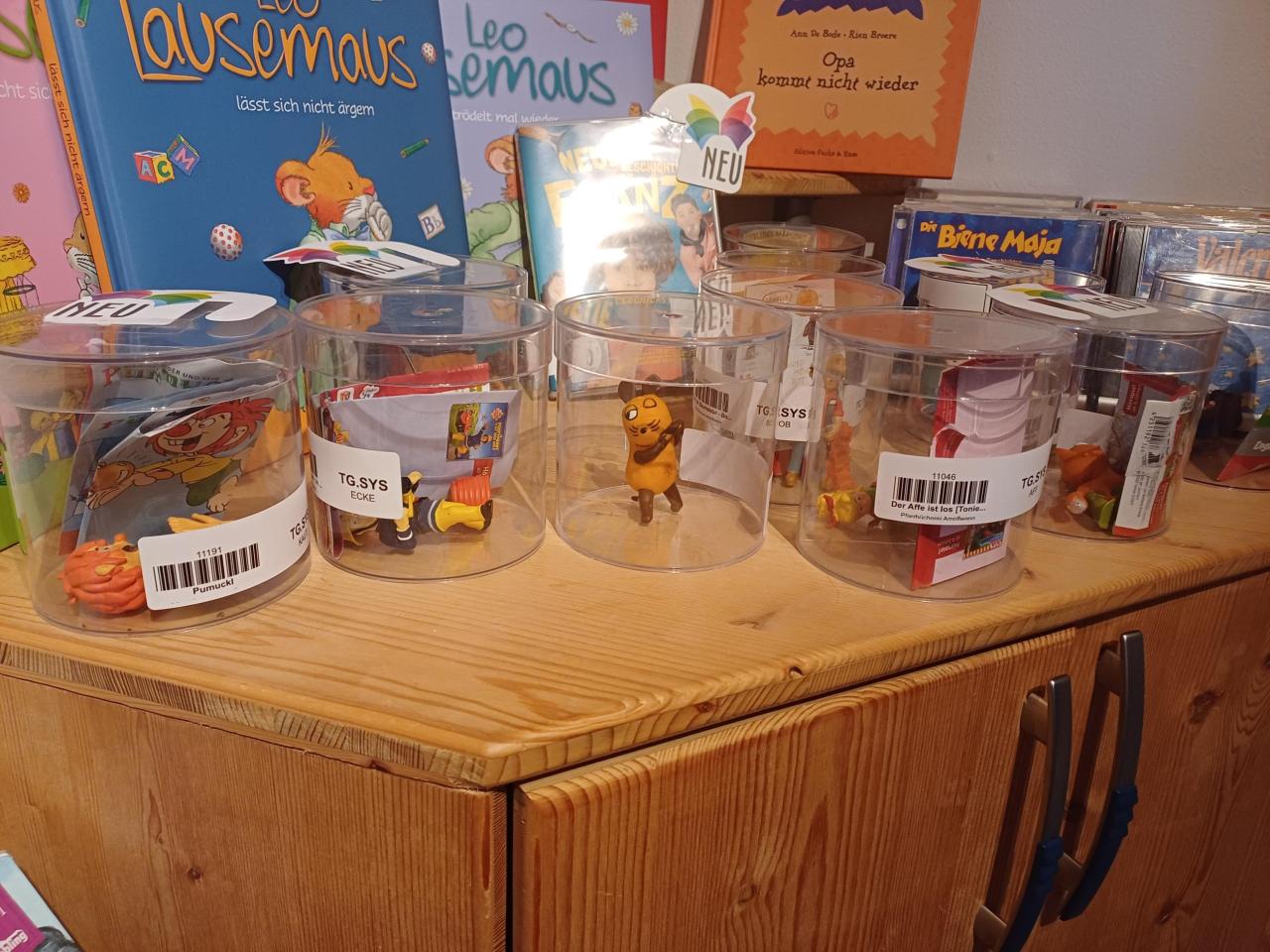 Spielzeugfiguren in transparenten Dosen auf einem Holztresen. Im Hintergrund stehen Kinderbücher wie "Lausemaus", "Leo Lausemaus" und "Opa kommt nicht wieder".