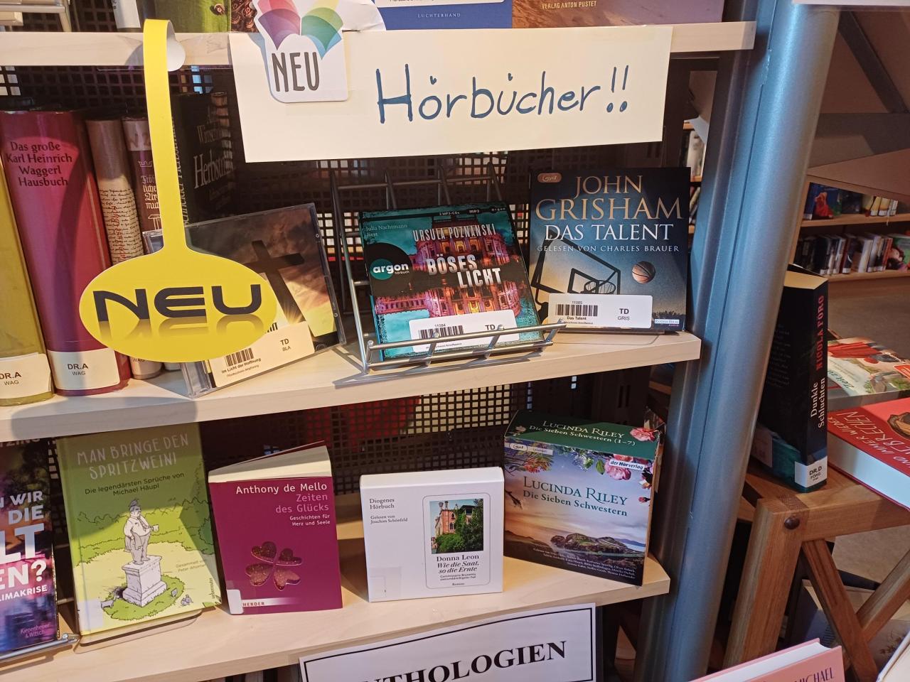 Regal mit neuen Hörbüchern in der Bibliothek. Sichtbar sind u. a. „Böses Licht“ von Ursula Poznanski und „Das Talent“ von John Grisham. Gelbes „NEU“-Schild und Hinweis „Hörbücher!!“.