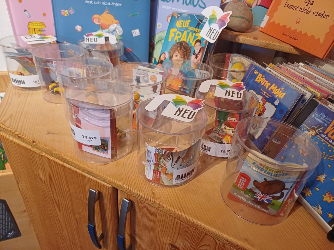 Reihe von durchsichtigen Dosen mit kleinen Spielzeugartikeln und "NEU"-Aufklebern auf einer Theke. Rechts liegen Stapel von Kinderhörspielen und CDs.
