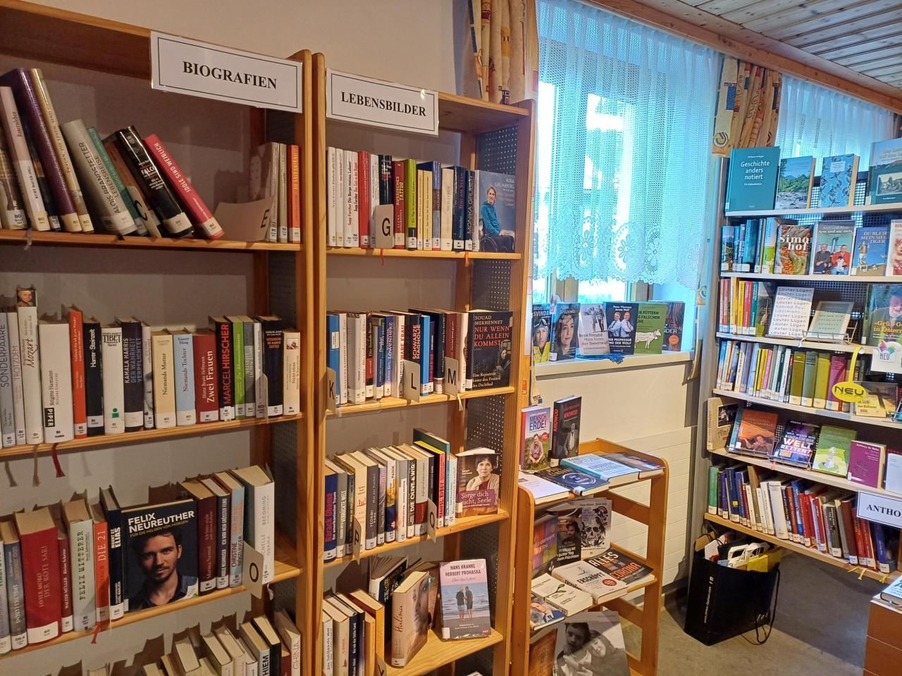 Holzregale, beschriftet mit "BIOGRAFIEN" und "LEBENSBILDER", gefüllt mit Büchern. Rechts ein Fenster mit hellblauem Vorhang und weiteren Bücherregalen.