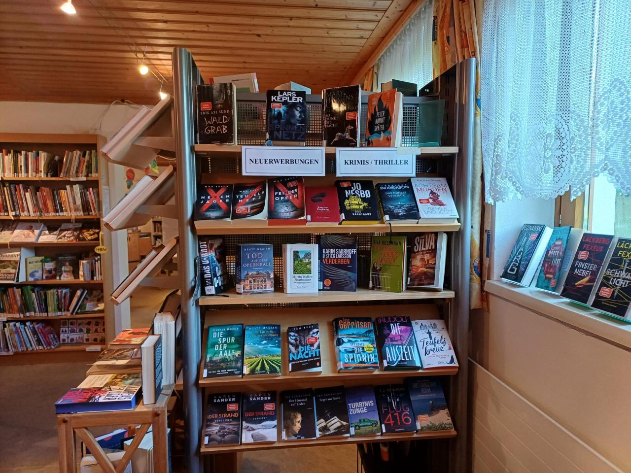 Mobiles Regal vor einem Fenster mit Spitze. Die Fächer sind mit neuen Krimis/Thrillern bestückt, beschriftet als "Neuerwerbungen" und "Krimis / Thriller".