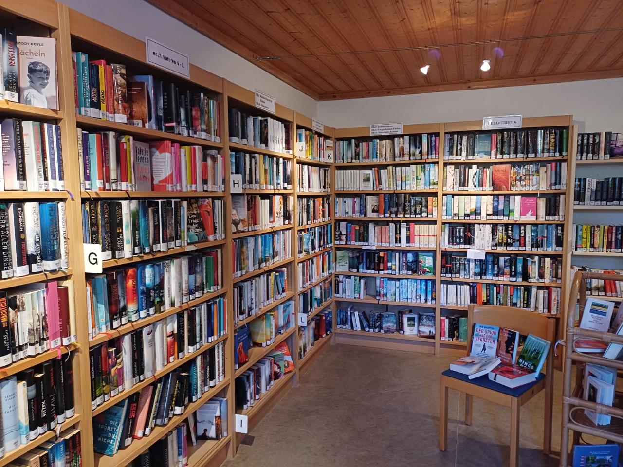Ecke einer Bibliothek mit hohen, mit Büchern gefüllten Holzregalen, die nach Buchstaben (G, H, I, J, K) und Themen ("Belletristik") sortiert sind.