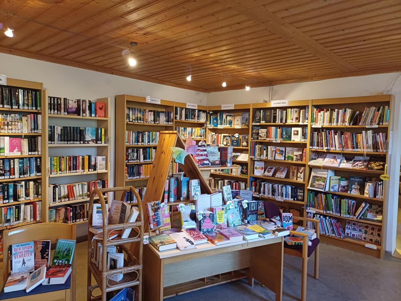 Überblick einer hellen Buchhandlung oder Bibliotheksecke mit Holzboden und -decke, umgeben von vollen Bücherregalen und einem zentralen Büchertisch.