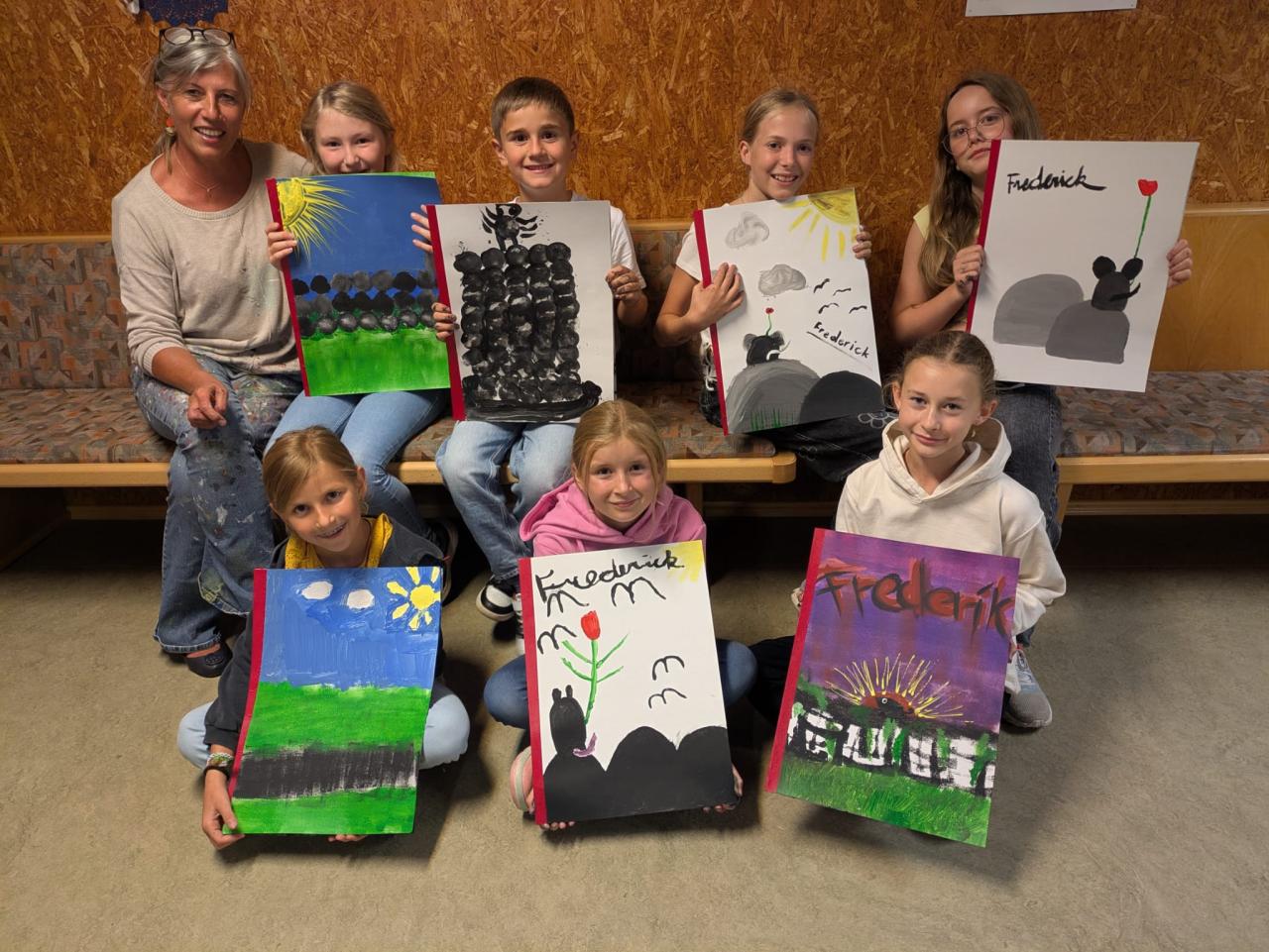 Sieben Kinder sitzen auf einer Bank und halten stolz ihre selbstgemalten Bilder hoch. Die bunten Kunstwerke zeigen Szenen aus der Geschichte „Frederick“ mit Mäusen, Blumen, Sonnenstrahlen und bunten Landschaften.