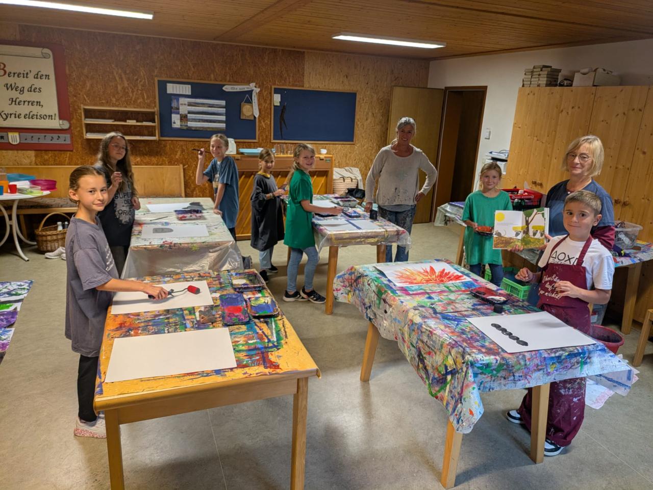 Kinder und zwei Erwachsene stehen an bemalten Tischen in einem Bastelraum. Die Kinder malen gerade Bilder zur Geschichte „Frederick“, einige arbeiten schon mit Farbe, andere zeigen ihre fertigen Werke.
