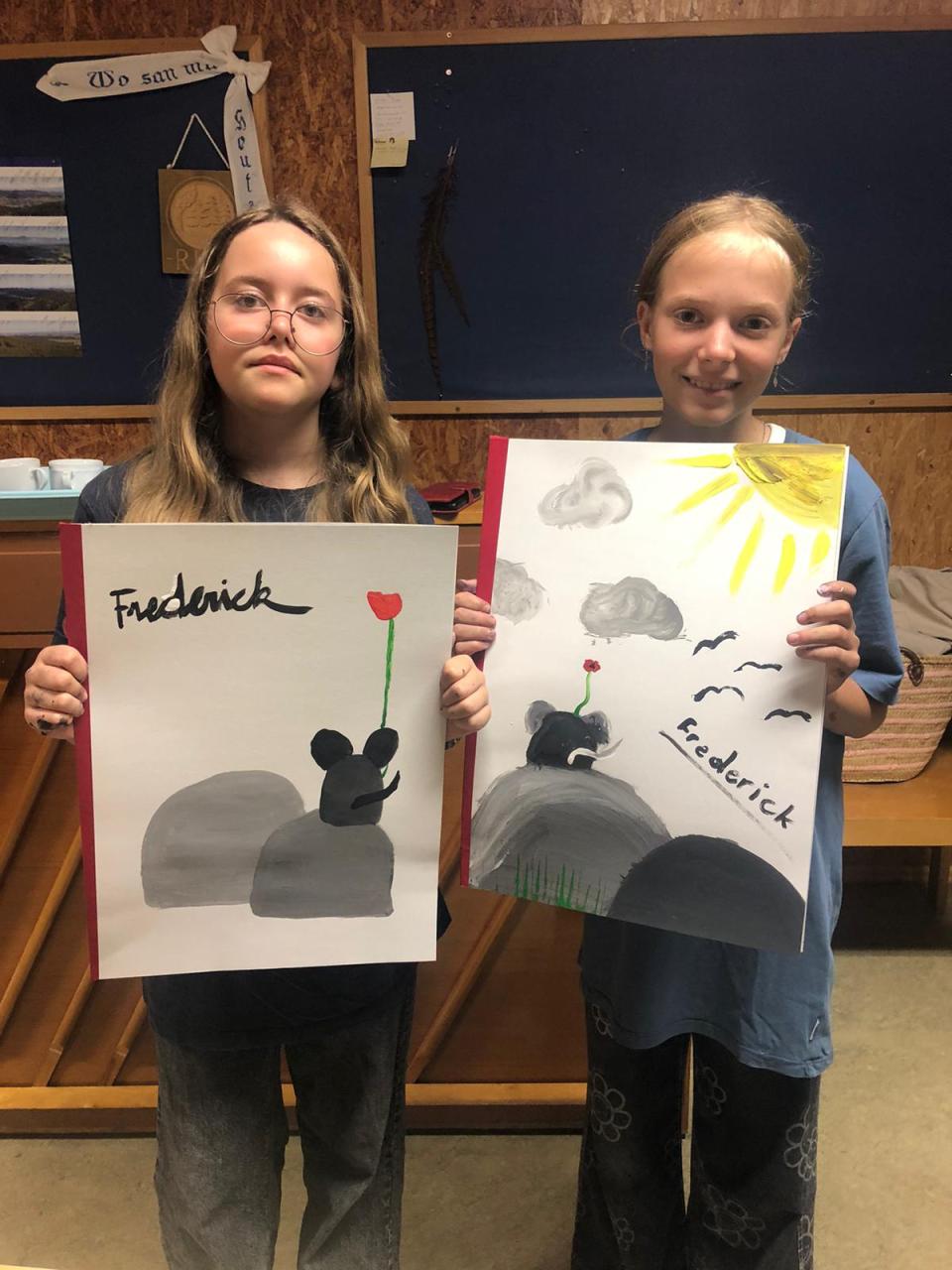 Zwei Mädchen halten stolz ihre Kunstwerke in die Kamera. Beide Kunstwerke zeigen eine Maus auf einem Felsen mit einer Blume, und auf jedem Bild steht „Frederick“, der Titel eines Kinderbuchs. Die Mädchen stehen in einem Raum mit einem Klavier im Hintergrund. Sie sehen glücklich und zufrieden mit ihren Bildern aus.