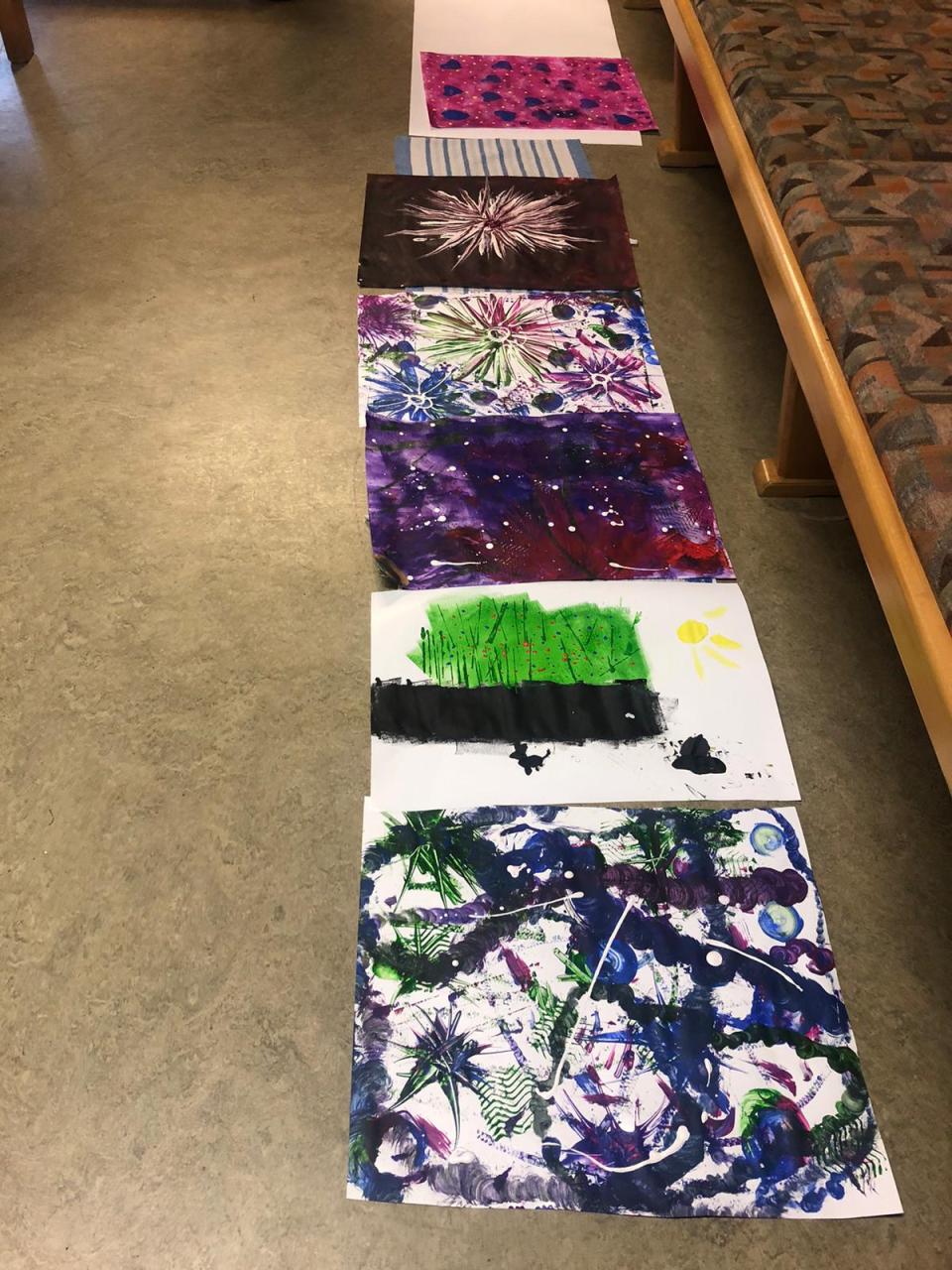 Verschiedene Kunstwerke, die auf dem Boden in einer Reihe liegen und von links nach rechts einen Weg bilden. Die Bilder sind in verschiedenen leuchtenden Farben und zeigen abstrakte Muster. Jedes Kunstwerk ist einzigartig und kreativ gestaltet. Die Werke sind auf dem Boden eines Innenraums ausgelegt.