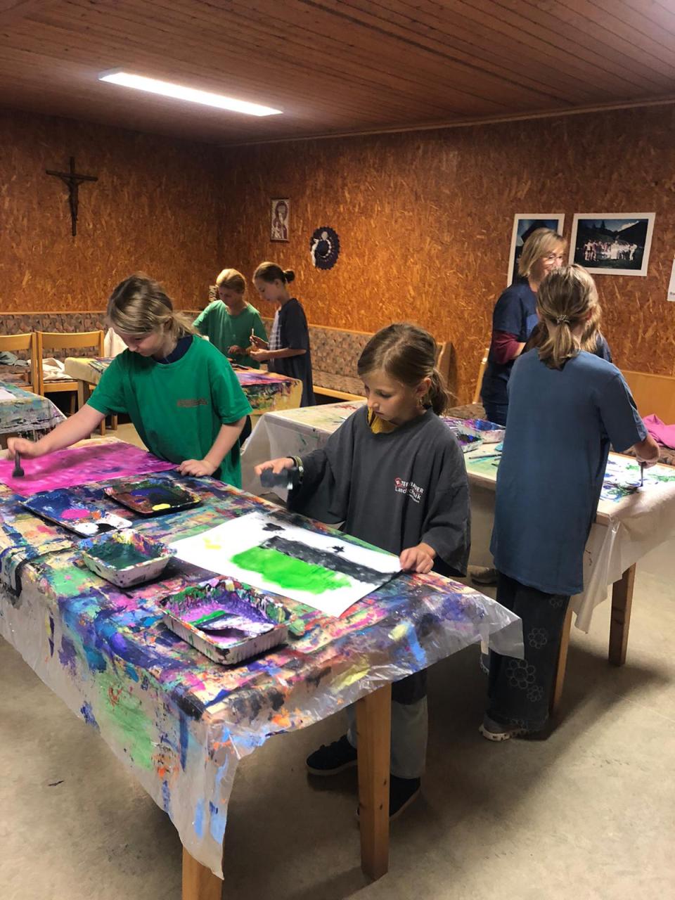 Kinder und Erwachsene sind in einem Atelier oder Kunstraum bei der Malerei zu sehen. Die Tische sind mit bunten Farben und Kunstmaterialien gefüllt. Ein Mädchen trägt eine Schürze und ist auf ihr Bild konzentriert. Die gesamte Szene ist lebendig und zeigt eine Kunstklasse in Aktion.