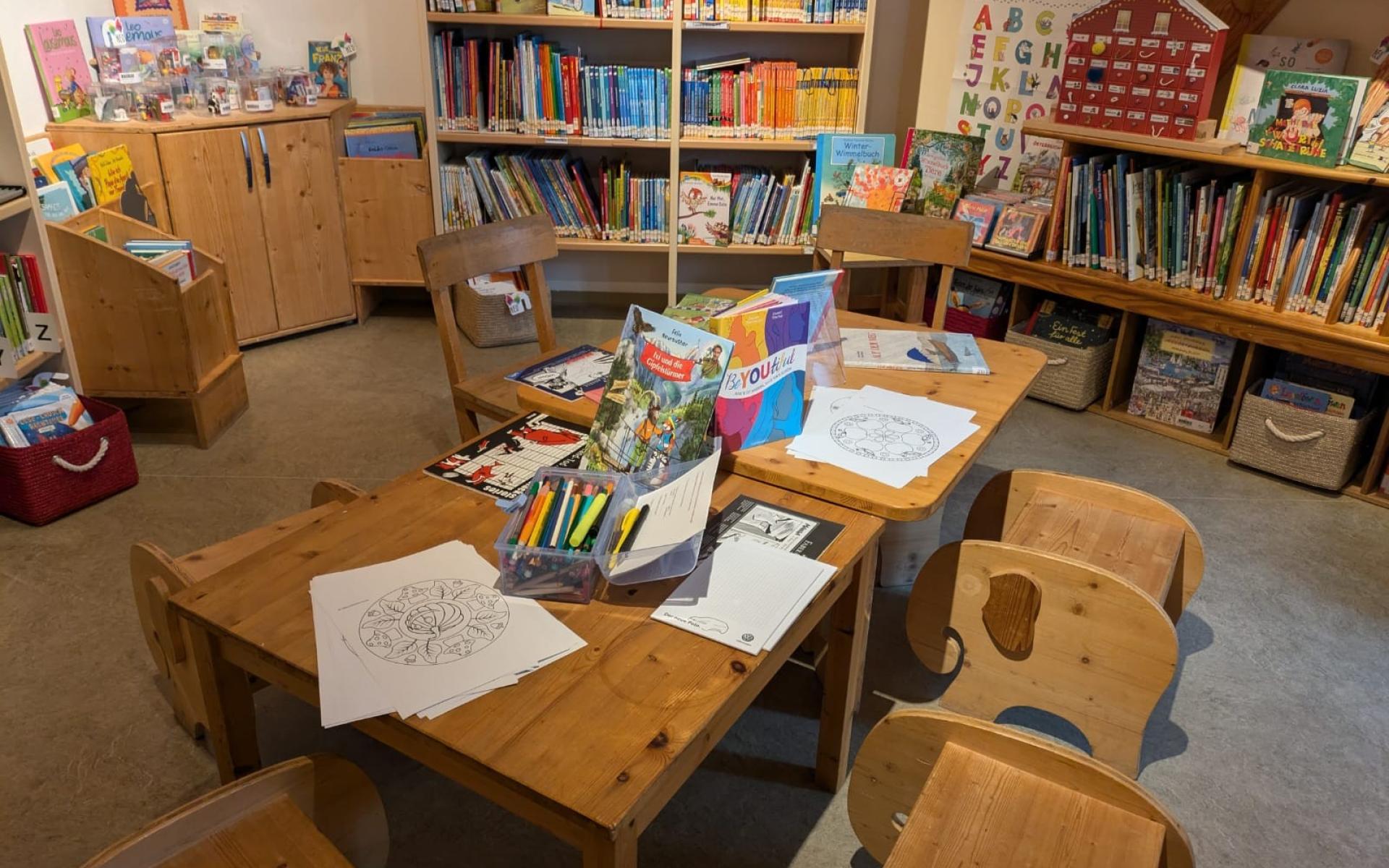 Kinderbereich einer Bibliothek mit niedrigen Holztischen und -stühlen, umgeben von Regalen voller bunter Kinderbücher; auf dem Tisch liegen Ausmalbilder, Buntstifte und Bücher bereit.
