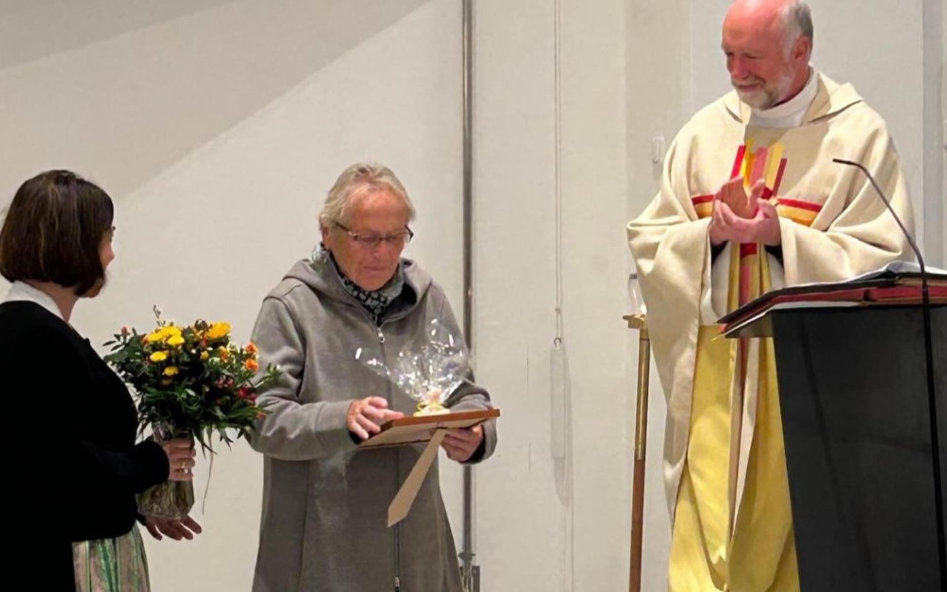 Renate Fellinger erhält bei einer Feier in der Kirche eine Urkunde und ein Geschenk, daneben eine Frau mit Blumen und ein Priester am Rednerpult.