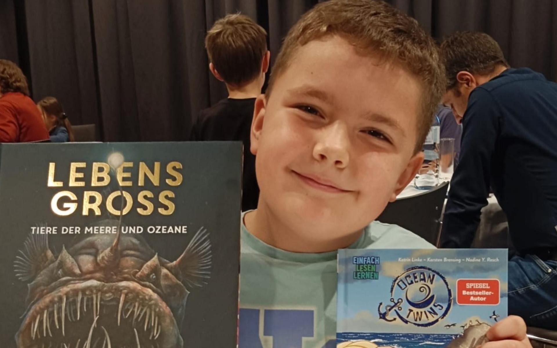 Junge sitzt an einem Tisch und hält stolz zwei Bücher in die Kamera – „Lebensgroß – Tiere der Meere und Ozeane“ und „Dieb-Alarm!“.