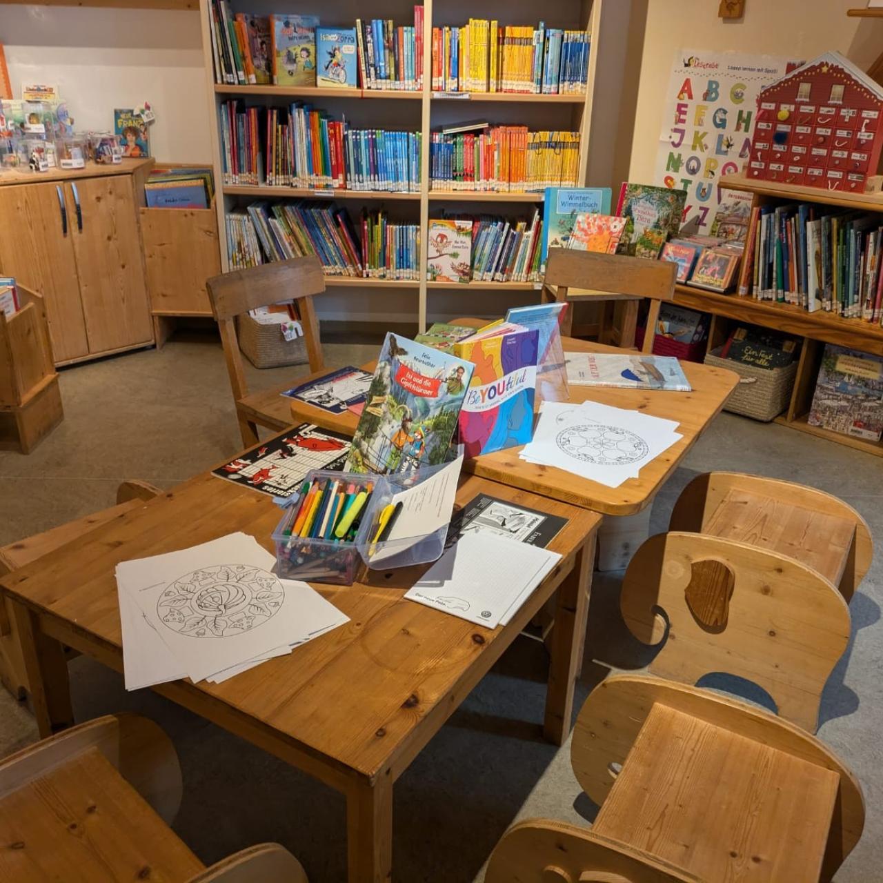 Kinderbereich einer Bibliothek mit niedrigen Holztischen und -stühlen, umgeben von Regalen voller bunter Kinderbücher; auf dem Tisch liegen Ausmalbilder, Buntstifte und Bücher bereit.